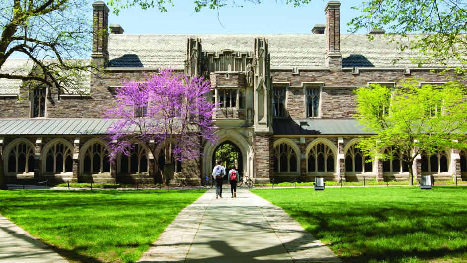 Two students on a Holder Hall cloister pathway surrounded by green grass and stone architecture
